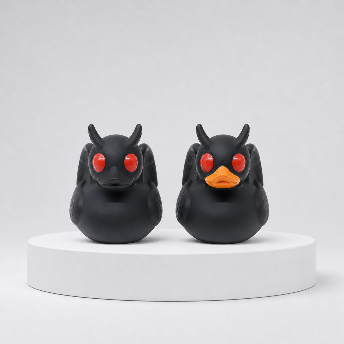 Two Mothduck cryptid collectibles shown together, featuring black bodies, red eyes, horns and wings with a choice of black or orange bill.