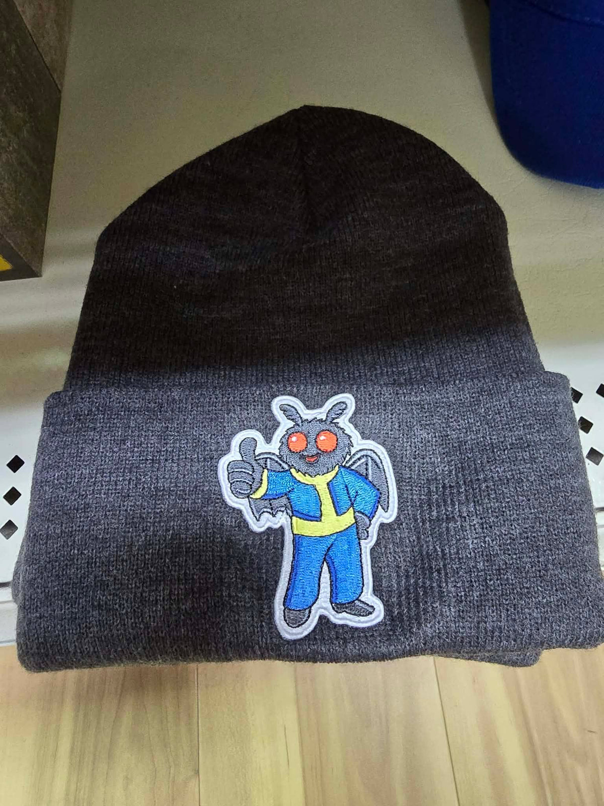 Charcoal gray cuffed knit beanie with embroidered Vault Guardian Mothman patch featuring glowing red eyes and a blue-and-yellow vault suit.
